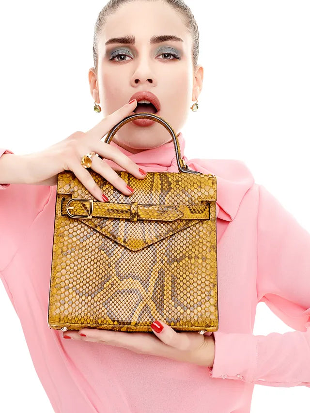 Model in a pink blouse holding a mustard python-leather top-handle bag to her lips