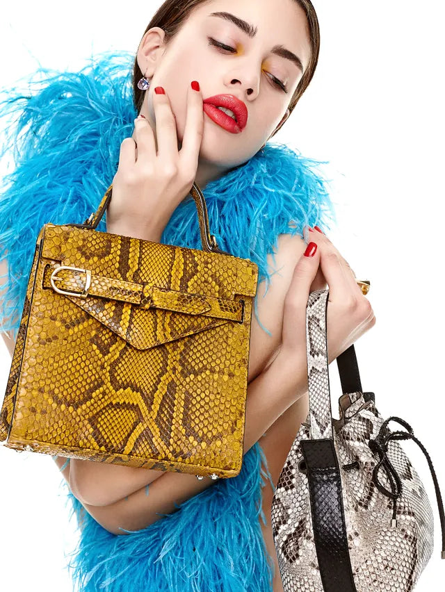 Model in a blue feather boa holding a mustard python-leather handbag and a python bucket bag