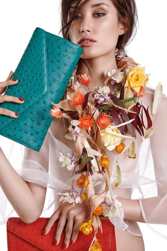 Model holding a teal ostrich-leather clutch beside a cascade of dried flowers, with a red clutch below