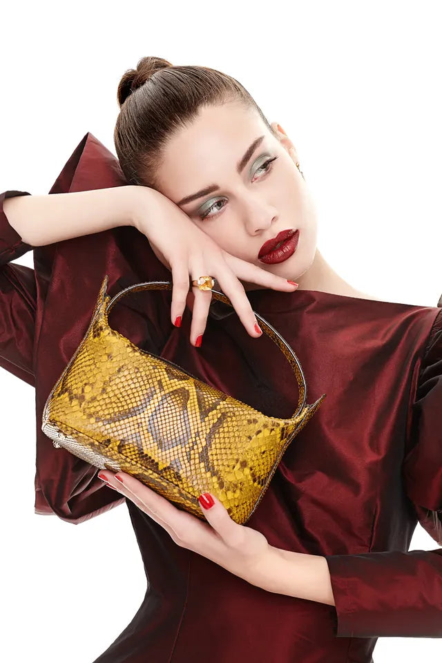Model in a burgundy silk dress cradling a mustard python-leather shoulder bag